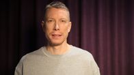 SDP:n kansanedustaja, psykoterapeutti Ville Merinen sinertävien verhojen edessä seisten kameraan katsoen YLE:n tiloissa.