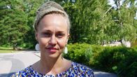 A woman with blonde dreadlocks, white earrings and a blue-pattern dress stands outdoors in a summer suburban setting.
