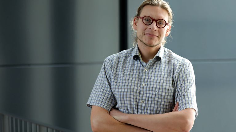 Ruutupaitaan pukeutuneella miehellä on tummanpunaiset pyöreät silmälasit. 