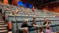 Auditoriossa noin viisikymmentä ihmistä yleisötilaisuudessa. Keskellä punapaitainen nainen on noussut ylös ja lukee asiaa paperista.