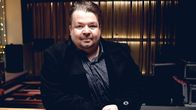 Tummiin pukeutunut keski-ikäinen mies. Tumma tausta, mahdollisesti studio tai bändin harjoittelutila.
