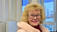 Vaaleahiuksinen, silmälasipäinen nainen hymyilee ikkunan edessä sisätiloissa.