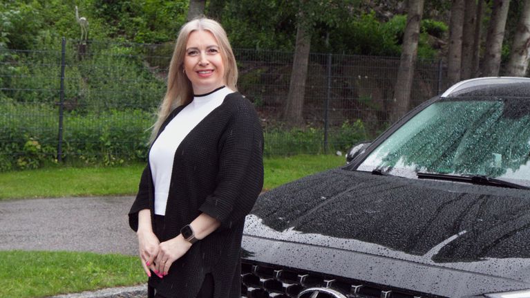 En kvinnlig taxichaufför står framför sin bil och ler i regnet.