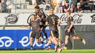Fyra fotbollsspelare i svarta matchkläder firar på en gräsplan framför reklamskyltar.