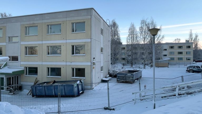 Vanha, betonirakenteinen, kolmikerroksinen kerrostalorakennus, jonka edessä on jätelavoja. Rakennus on aidattu metalliaidalla. 