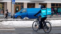 Woltin ruokalähetti ylittää lumista tietä polkupyörällä. Taustalla jalankulkija ja pakettiauto.