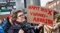 Nainen seisoo kadulla kädessään pahvikyltti, jossa lukee “NÄPIT IRTI X VIIANKiAAVASTA”, taustalla muita ihmisiä.