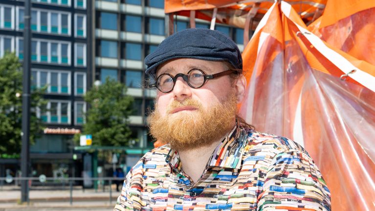 Pauli Rautiainen istuu pienen värikkään pöydän ääressä. Taustalla on torikojun oranssi pressu ja hotellirakennus.