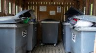 Sekajätteen keräyslaatikoiden kannet eivät mene kiinni. Raollaan ja osittain auki olevien kansien alta näkyy kirkkaita, vihreitä, mustia ja kirjavia muovipusseja, joissa on roskia. Kopin takaseinälle on kiinnitetty valkoisia lappuja. 