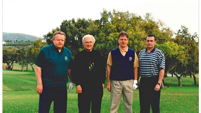 Neljä miehen ryhmä seisoo viheriöllä golf-kentällä, taustalla puita ja vihreää nurmikkoa.  