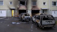 Kerrostalon alakerroksessa kaksi palanutta autoa ja ikkunoissa nokijälkiä; etualalla vaahtojäämiä asfaltilla.