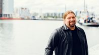 En man står i en hamn med suddiga båtar och hus i bakgrunden.
