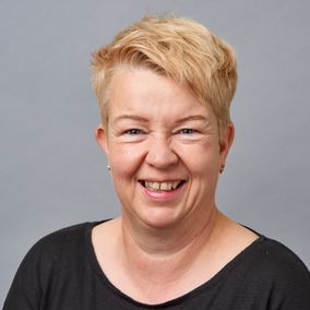 Vaaleahiuksinen, keski-ikäinen nainen katsoo kameraan ja hymyilee.