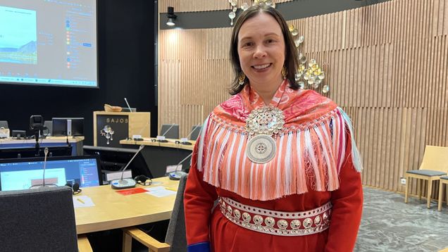 Nainen saamelaispuvussa seisoo kokoushuoneessa, taustalla näkyy Sajos-teksti sekä tietokonenäyttöjä 24.9.2026 Inarissa.