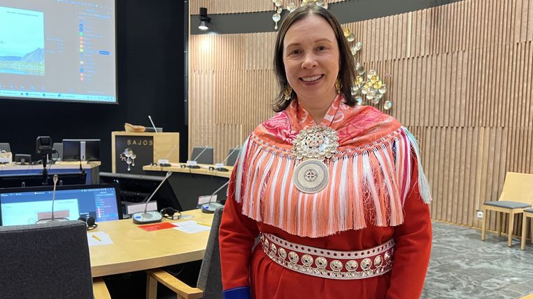 Nainen saamelaispuvussa seisoo kokoushuoneessa, taustalla näkyy Sajos-teksti sekä tietokonenäyttöjä 24.9.2026 Inarissa.