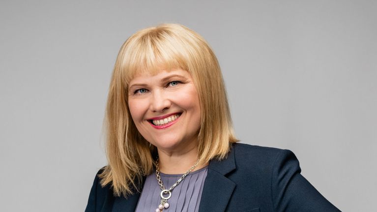 Vaaleahiuksinen nainen hymyilee kameralle. Hänellä on päällään harmaa paita ja jakkupuku.