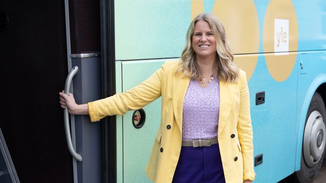 En person står framför en blå och grön buss med texten ”100 yle” på sidan, en stol står bredvid bussen.