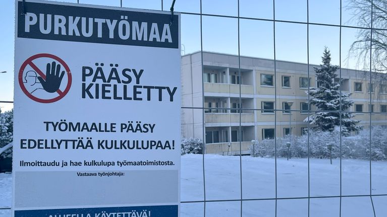 Kyltti metalliaidassa. Kyltissä lukee: purkutyömaa, pääsy kielletty. Taustalla näkyy vanha, betonirakenteinen, kolmikerroksinen kerrostalorakennus. 