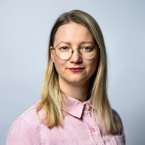 Henkilö pukeutunut vaaleanpunaiseen kauluspaitaan seisoo vaalealla, neutraalilla taustalla. Hänellä on keskipitkät vaaleat hiukset.