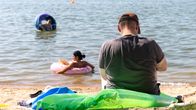 En man sitter på stranden på en luftmadrass. I havet simmar en kvinna och ett barn i simringar.