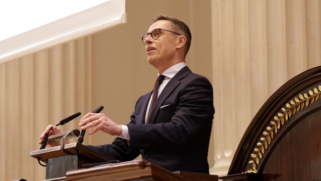 Mies seisoo puhujakorokkeella suuressa salissa ja puhuu mikrofoneihin; taustalla näkyy vaalea seinä ja pylväitä.
