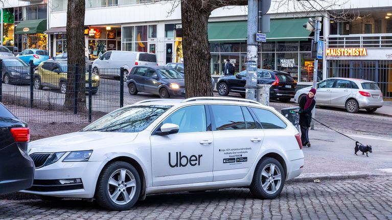 A white estate wagon with the Uber logo on its side, parked in a city with trees and a dogwalker behind it.