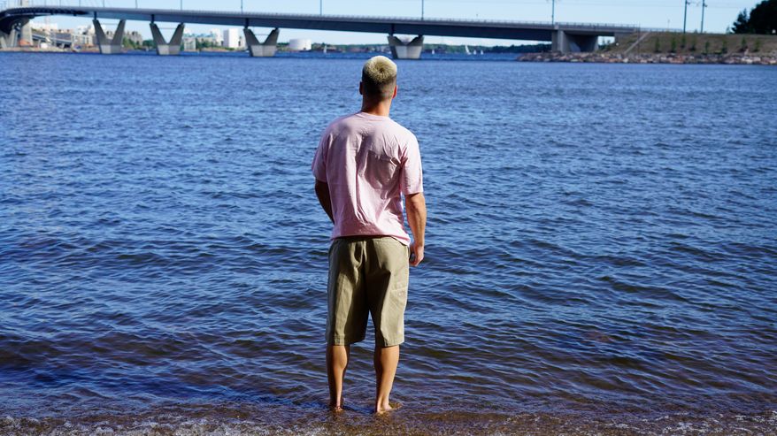 Mies seisoo selin rantavedessä ja katsoo horisontissa näkyvää siltaa.