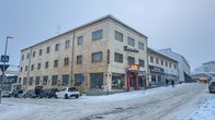 Kuvassa on talvinen kaupunkimaisema, jossa näkyy kulmikas kerrostalo. Rakennuksessa on liiketiloja alimmassa kerroksessa.