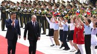Xi Jinping ja Vladimir Putin kävelevät punaista mattoa pitkin vierekkäin ja vilkuttavat Venäjän ja Kiinan lippuja heiluttaville lapsille. Lasten takana seisoo rivissä sotilassoittokunta.