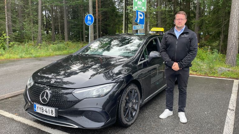 En man står bredvid en svart Mercedes-taxi på en parkeringsplats.
