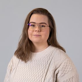 Henkilö pukeutunut beigeen neulepaitaan poseeraa harmaata taustaa vasten.