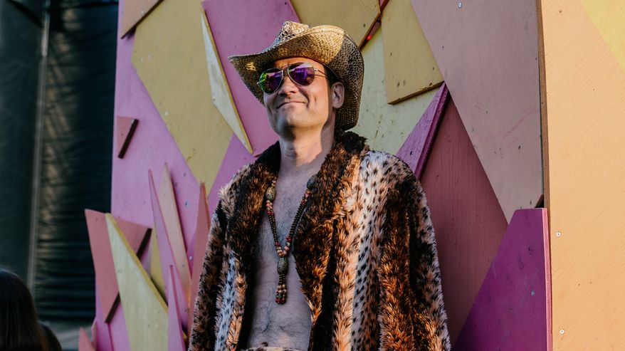 Mies poseeraa värikkään taustan edessä pukeutuneena leopardikuosiseen turkkiin ja punaiseen asusteeseen, cowboy-hattu ja valkoiset cowboy-bootsit täydentävät hänen asukokonaisuutensa.