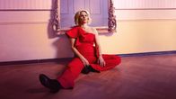 Linda (Laura Malmivaara) iklädd en röd elegant byxdress sitter på golvet och ser uppgiven ut.