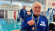 Harmaahiuksinen mies esittelee kultamitalia urheiluhallissa. 
