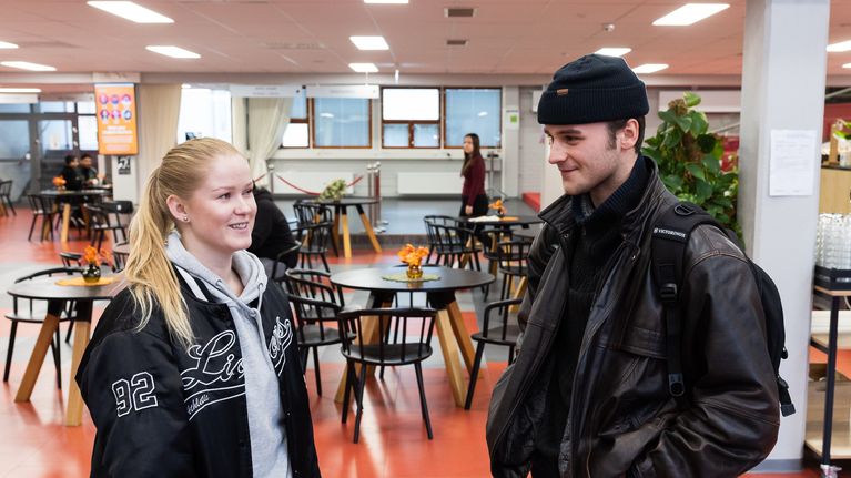 Nuori nainen ja nuori mies aulassa. Miehellä on reppu selässä ja naisella laukku olalla.