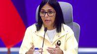 Venezuelas tillförordnade president Delcy Rodriguez iklädd glasögon och ljus jacka sitter vid ett bord med mikrofon, Venezuelas flagga i bakgrunden.