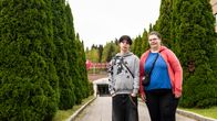 Kuvassa kaksi henkilöä seisoo asvaltoidulla pihalla, taustalla näkyy korkeita havupuita ja suuri rakennus.