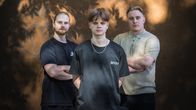 Niklas Mäkelä, Emil Haataja ja Heikki Kokkonen poseeraavat seinän edessä. Seinälle osuu auringonsäteitä ja puun varjo.