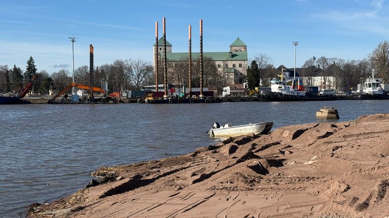 En strand med sandhögar vid vattnet, i bakgrunden ser man fartyg och Åbo slott.
