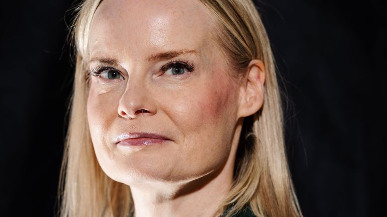 Riikka Purra i en mörkgrön kavaj och vit t-tröja tittar in mot kameran. Hon är fotograferad vid en svart vägg.