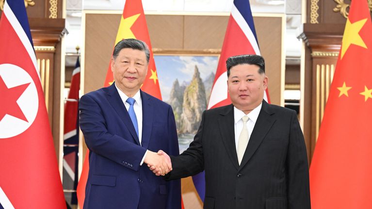 Kaksi pukupukuista miestä kättelee Pohjois-Korean ja Kiinan lippujen edessä juhlavassa sisätilassa. Taustalla on maisemamaalaus.