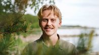 Nuori vaaleantukkainen mies hymyilee kameraa kohti meren rannalla. Männyn oksat kehystävät kasvoja. Aurinko paistaa. 