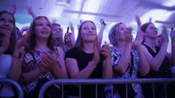 Eturivissä seisoo useita ihmisiä, jotka taputtavat käsiään valaistussa sisätilassa metalliaidan takana.