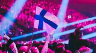 Suomen lippu liehuu yleisön joukossa areenalla, taustalla näkyy pinkkejä ja sinisiä valoja.