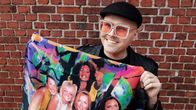 Eino Nurmisto seisoo punatiilisen seinän edessä. Hän katsoo kameraan hymyillen. Eino pitää käsissään Spice Girls–seinälippua. Hänellä on päässään musta hattu ja vaaleanpunaiset aurinkolasit, joissa on paljetteja sangoissa.