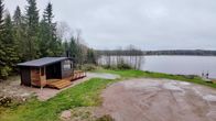 Saunarakennus puiden vieressä järven rannalla, nurmikkoa, laituri veden äärellä, pilvinen sää.