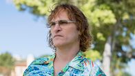 Näyttelijä Joaquin Phoenix puolilähikuvassa havaijipaidassa ja silmälaseissa, puolipitkä kihara tukka hapsottaen katsoo jonnekin kaukaisuuteen aurinkoisessa maisemassa. Kuva elokuvasta Ei hätää, hän ei kävele pitkälle.