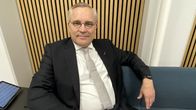 Silmälasipäinen, keski-ikäinen mies istuu tumma puku päällä sohvalla.