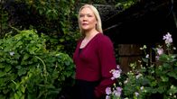 En kvinna med ljust hår står utomhus bland gröna växter och ljusrosa blommor, framför ett träskjul.