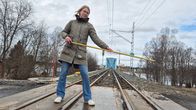 Toimittaja Laura Valta seisoo mittanauha kädessään lähellä junaraiteita. Taustalla näkyy Torniosta Ruotsiin kulkeva rautatiesilta.